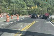 Vehicles moving through the N.C. 105 and Broadstone Road intersection on a green signal, with barricades closing the old bridge alignment at left.