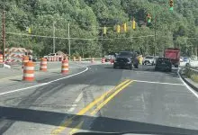 Vehicles moving through the N.C. 105 and Broadstone Road intersection on a green signal, with barricades closing the old bridge alignment at left.