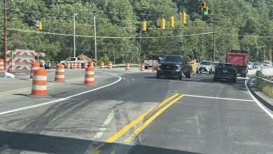 Vehicles moving through the N.C. 105 and Broadstone Road intersection on a green signal, with barricades closing the old bridge alignment at left.